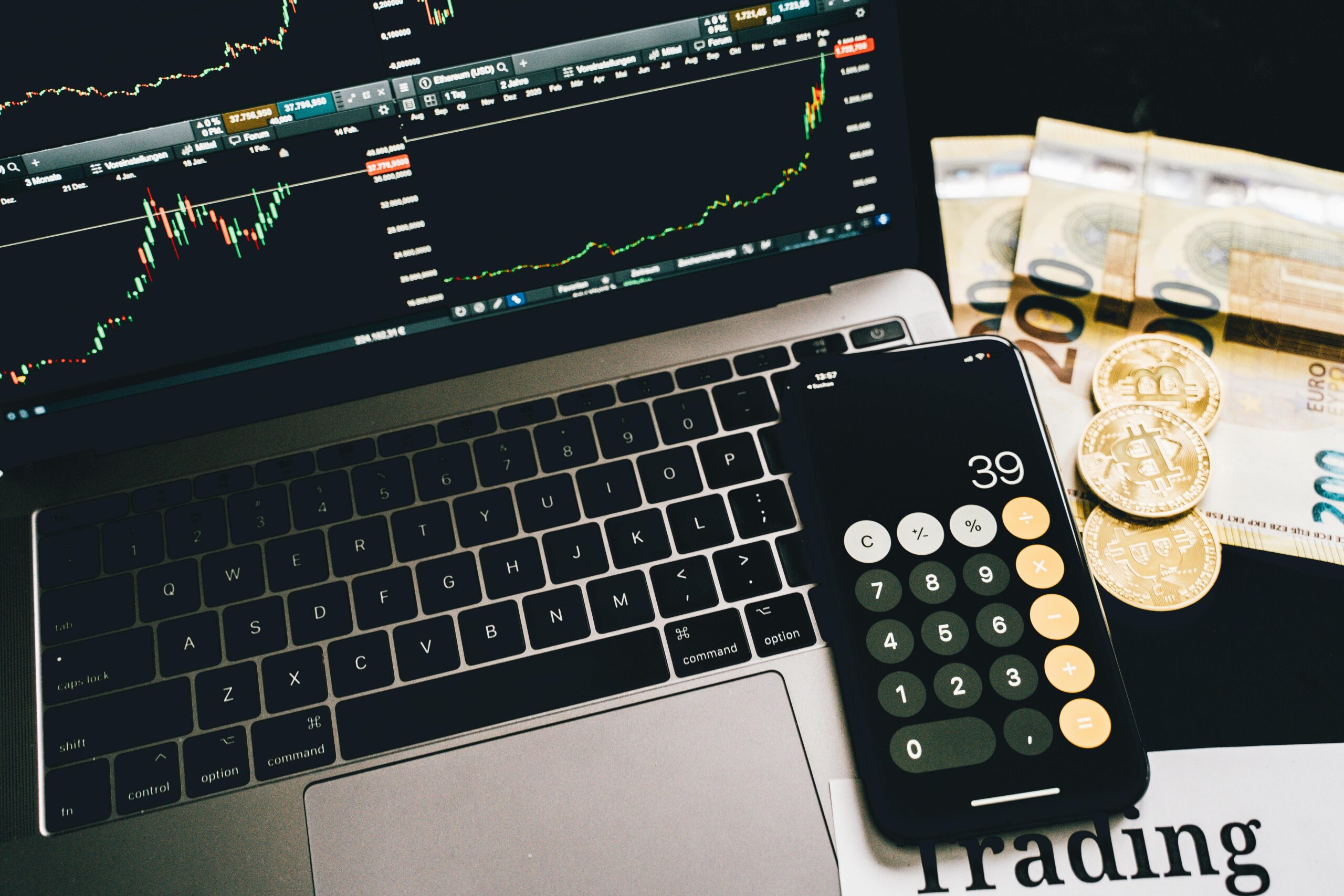 Laptop screen with trading charts, calculator app, Euros and Bitcoin coins on desk.